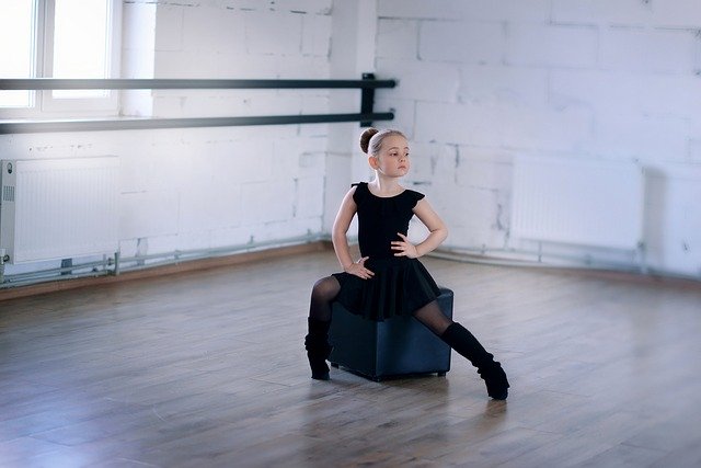 Menina pequena vestindo collant preto e polainas, sentada em um puff quadrado no centro de uma sala de dança.