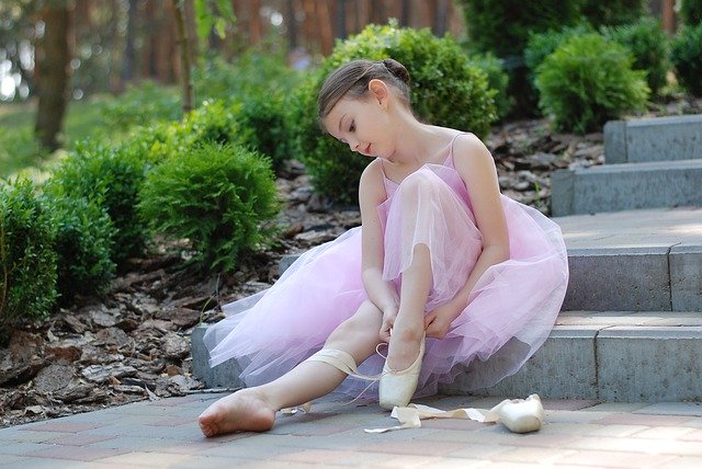 Menina pequena com roupas de ballet, sentada em uma escada, colocando sapatilha em um de seus pés.