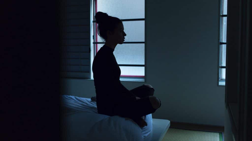 Mulher sentada na cama em um quarto escuro, pensativa.