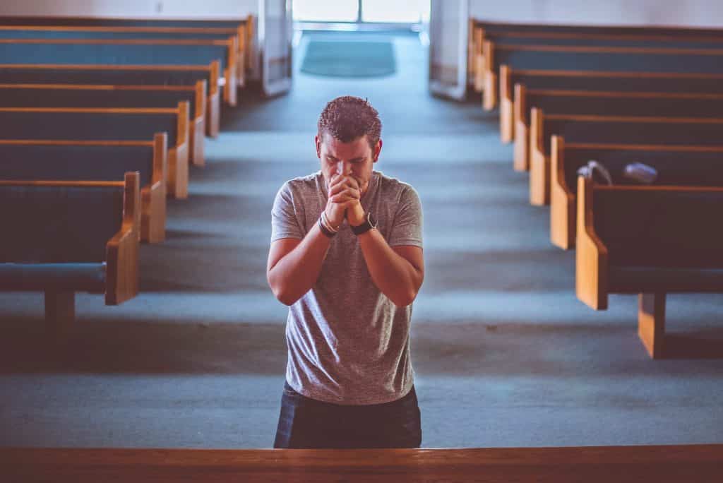 Homem em pé, com as mãos juntas sobre o rosto, orando em uma igreja.