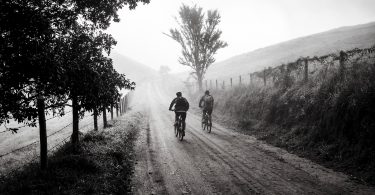 Imagem em preto e branco de uma estrada com um casal de amigos andando sobre ela de bicicleta.