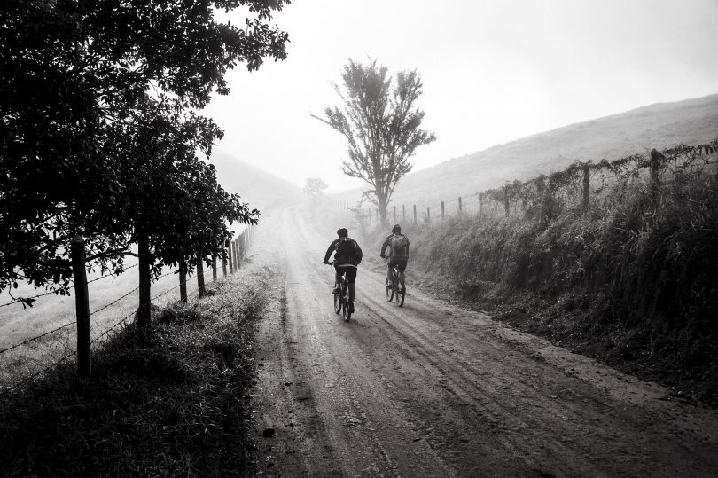 Imagem em preto e branco de uma estrada com um casal de amigos andando sobre ela de bicicleta.