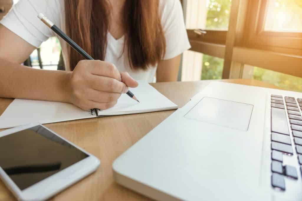 Mulher escrevendo com lápis em um caderno, com um celular ao lado e um notebook em sua frente.