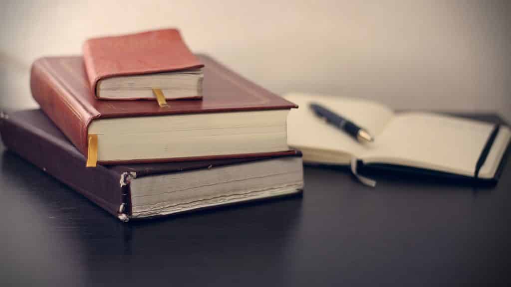 Imagem de três livros espíritas fechados um sobre o outro. O primeiro livro é o menor de todos.  Eles estão dispostos sobre ume mesa preta. Ao lado um caderno de anotações e uma caneta sobre ele.