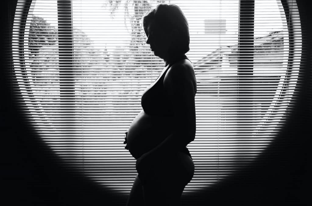 Mulher grávida de perfil em frente à uma janela.