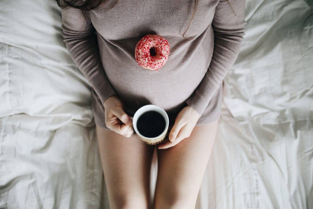 Mulher grávida sentada na cama com uma rosquinha apoiada na barriga e segurando uma xícara de café.