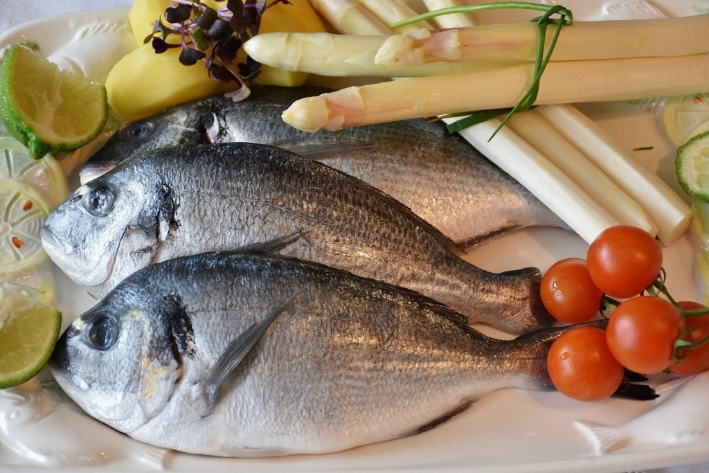 Imagem de três peixes crus separados para serem preparados em uma refeição. Temos ainda, tomates, limões, aspargos que farão parte da receita,
