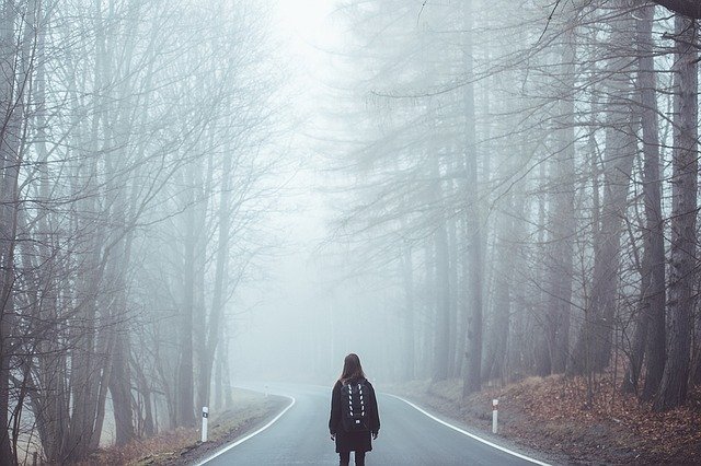 Mulher em pé no meio de rodovia com neblina e árvores dos dois lados