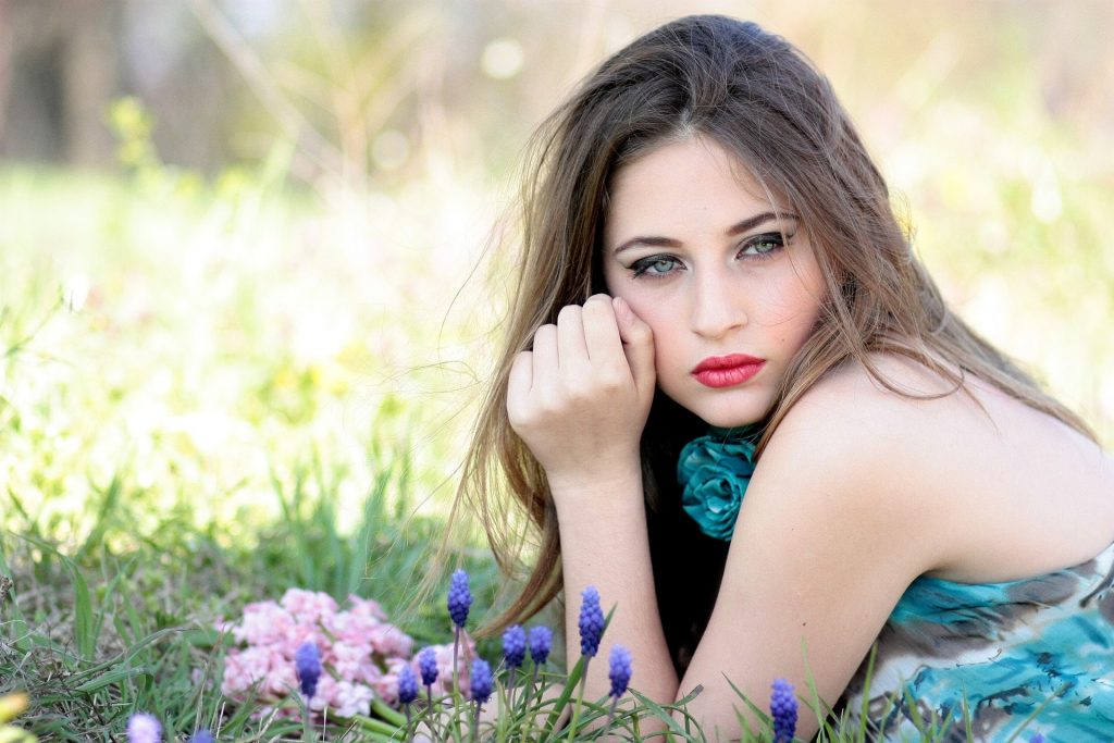 Imagem de uma linda mulher de olhos azuis, cabelos longos. Ela usa um batom vermelho e um vestido azul claro. Ela está deitada sobre um gramado com flores.