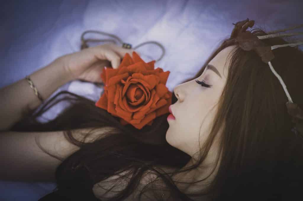 Imagem de uma mulher deitada sobre uma cama forrada com lençol branco. Ela está segurando um rosa vermelha em uma de suas mãos.