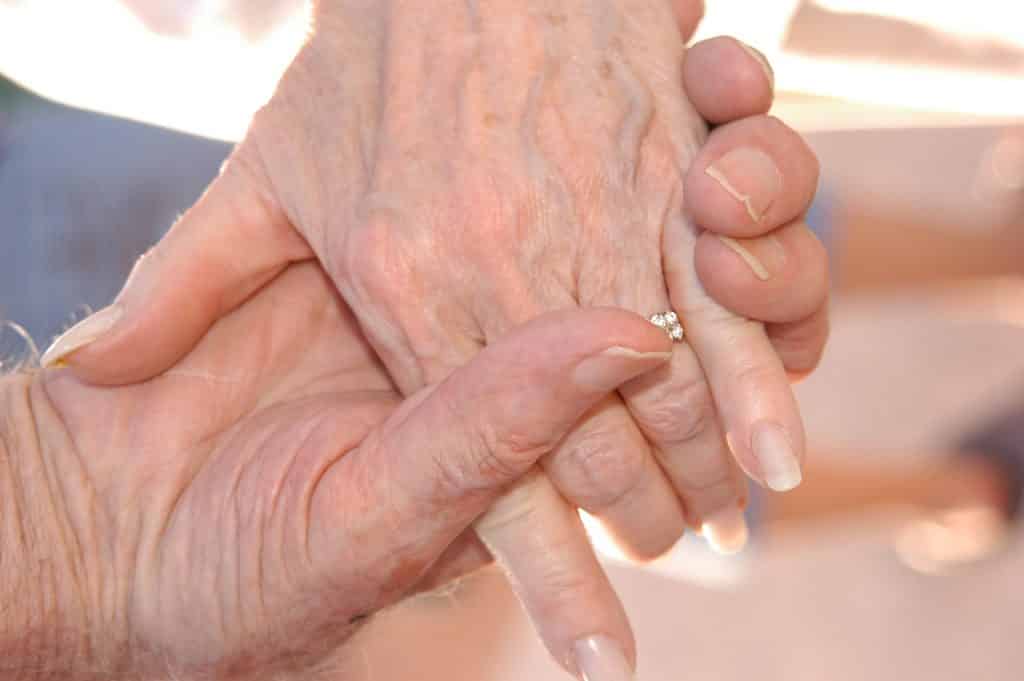 Imagem de duas mãos de pessoas idosas. A mão de um senhor segurando a mão de sua senhora.