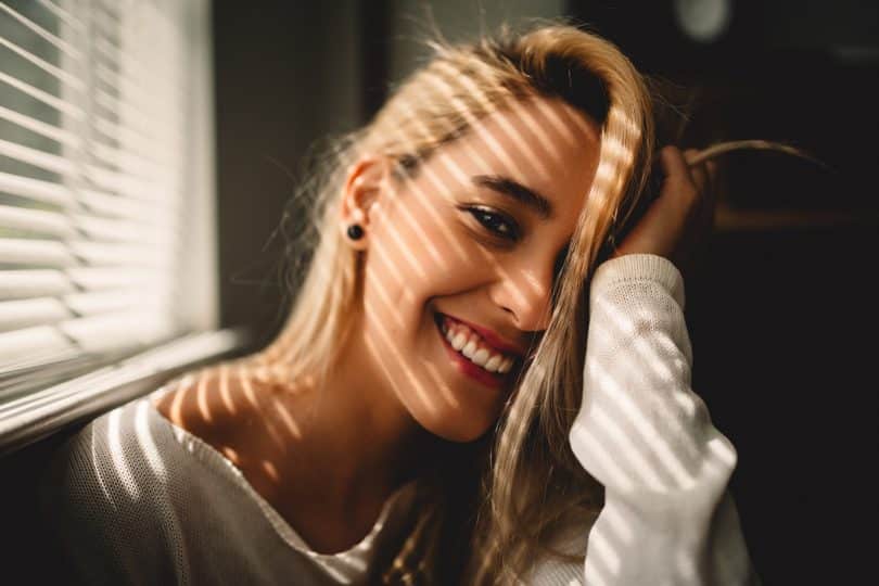 Mulher sorrindo apoiando sua mão na cabeça ao lado de uma janela