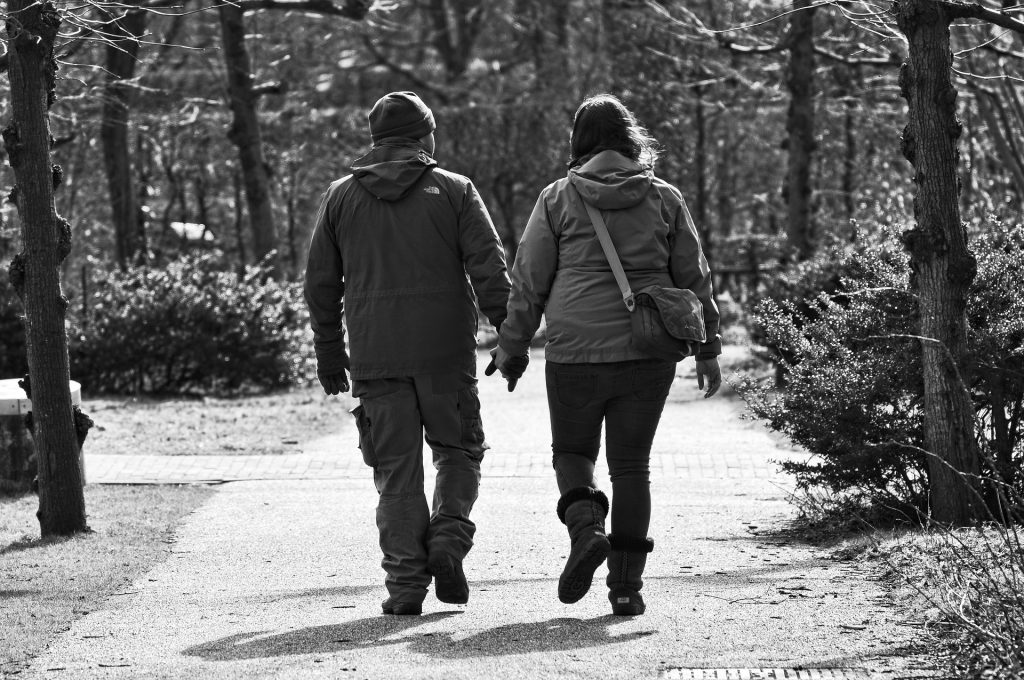 Imagem de um casal de meia idade vestidos com casados de frio. Eles estão de mãos dadas e caminham em um parque.
