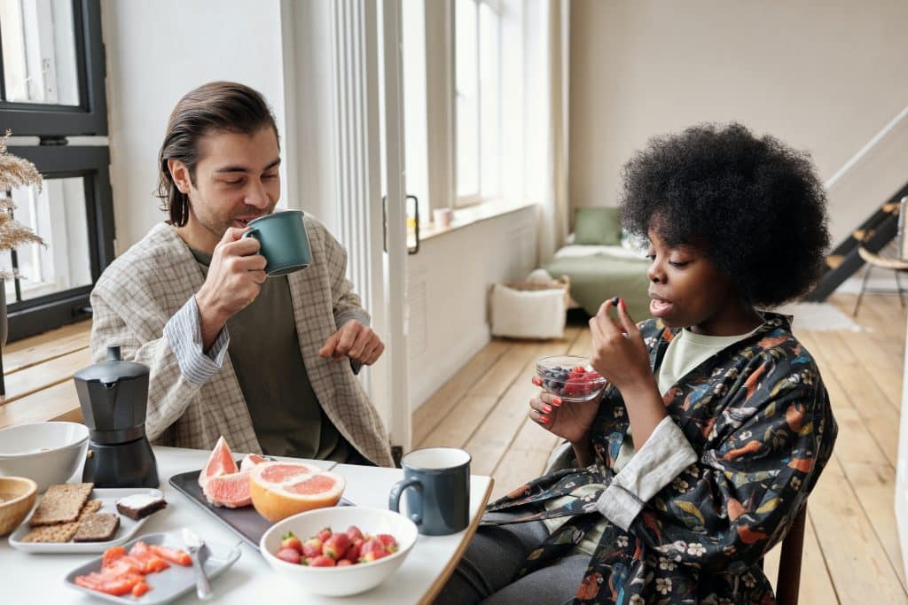 Homem e mulher sentados tomando café da manhã em uma mesa com frutas e canecas.