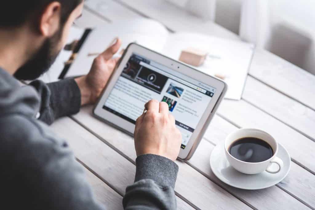 Homem estuda segurando um tablet sobre uma mesa com livros e uma xícara de café.