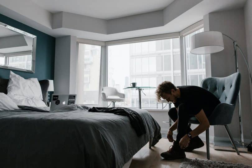 Homem em um quarto arrumado durante o dia, sentado em uma cadeira, amarrando os cadarços de sua bota.