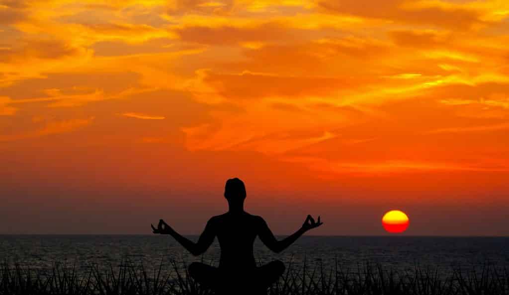 Imagem da silhueta de um homem meditando sentando de frente para o mar e ao fundo um lindo por do sol.