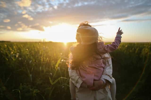 Mãe com filha nas costas olhando campo verde e sol refletindo ao fundo