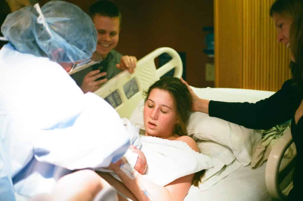 Mulher deitada em uma cama de hospital dando à luz ao filho.
