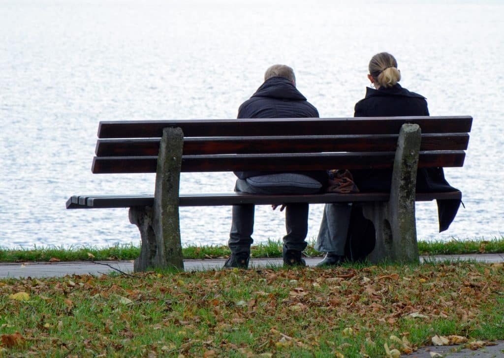 Imagem de um casal sentando em um banco de mandeira em frente a um lago. Ambos vestem casacos de inverno na cor azul.