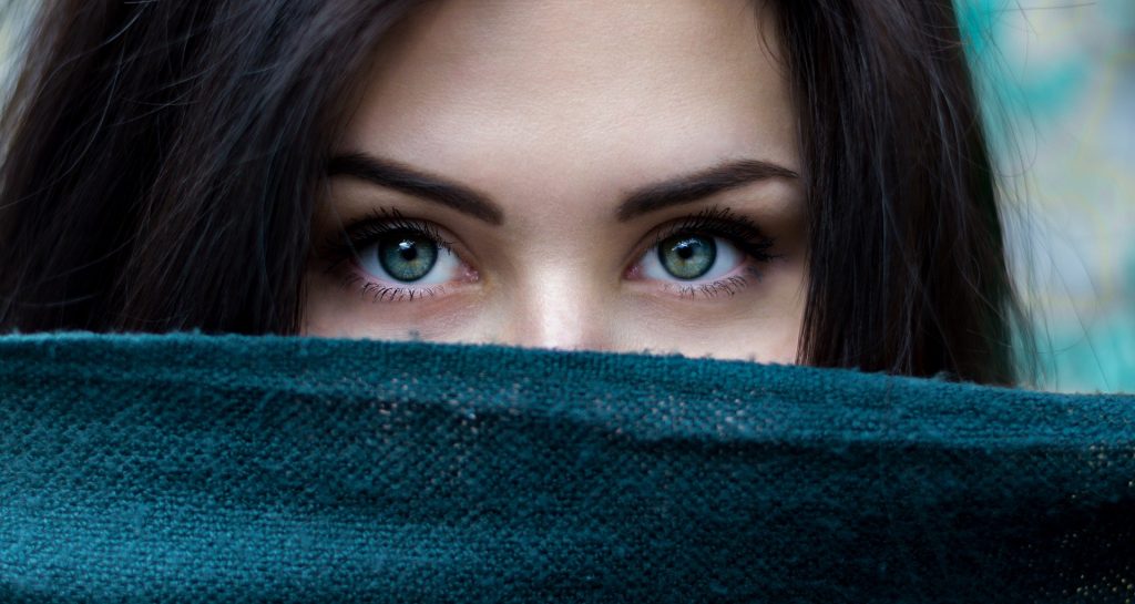Imagem dos olhos verdes de uma linda mulher de cabelos longos escuros. Ela segura um lenço verde que cobre o seu nariz e boca.