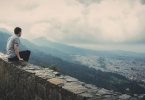 Homem sentado em muro baixo no topo de uma montanha, com vista para uma cidade.