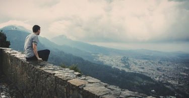 Homem sentado em muro baixo no topo de uma montanha, com vista para uma cidade.