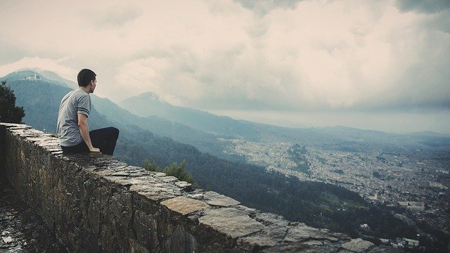 Homem sentado em muro baixo no topo de uma montanha, com vista para uma cidade.