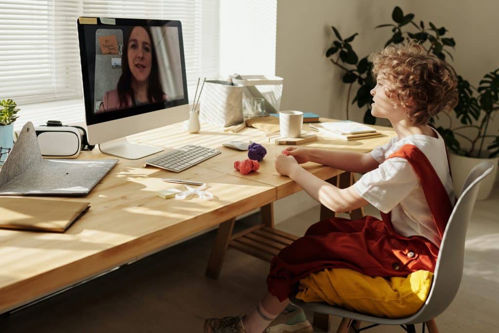 Criança sentada em frente a um computador, assistindo a uma aula por videochamada.