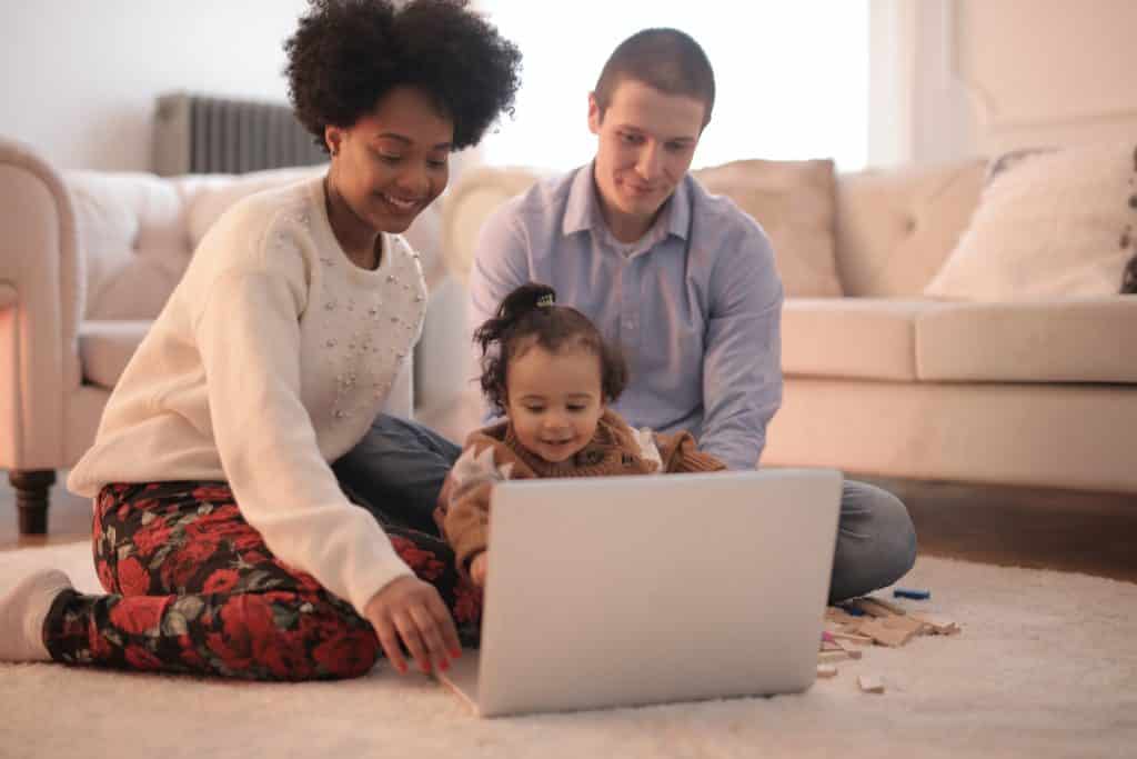 Homem, mulher e uma criança pequena sentados no chão da sala de estar, usando o computador.