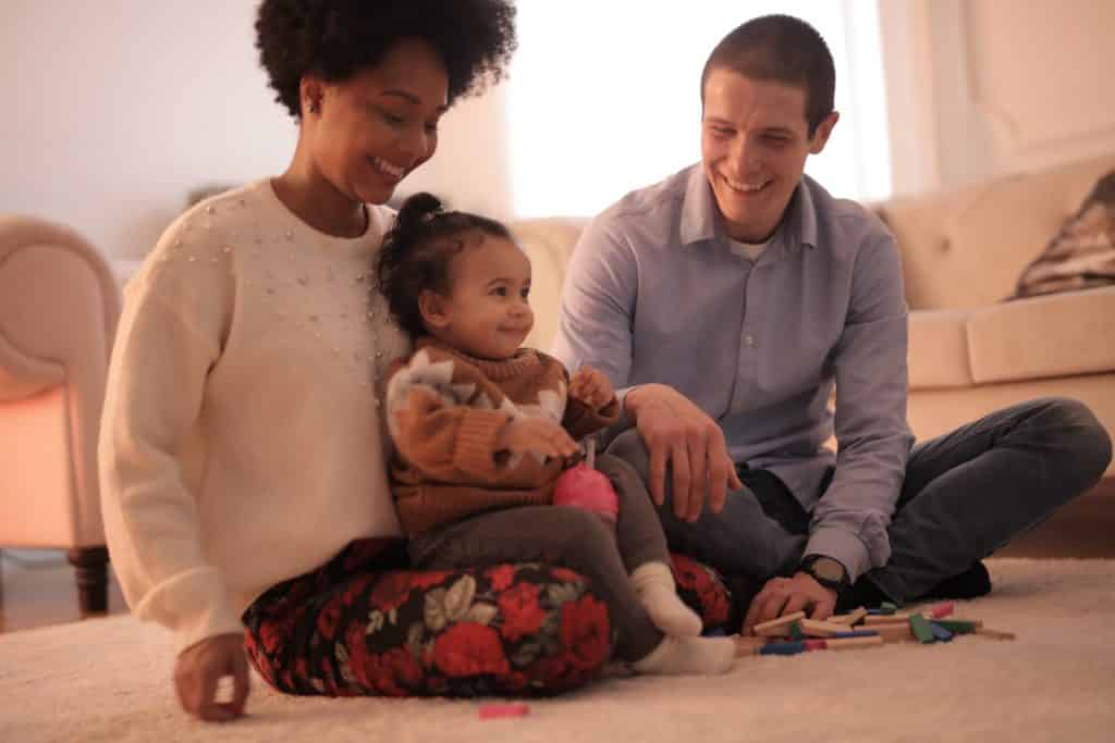 Homem e mulher sentados no chão, sorrindo. No colo da mulher, uma bebê também sorri.