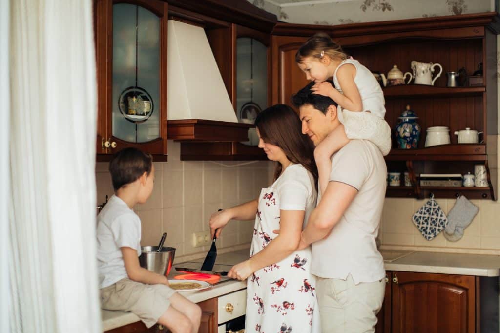 Família em cozinha. Um menino está sentado em uma bancada, uma mulher usa uma espátula dentro de uma frigideira e um homem segura uma menina pequena em seus ombros.