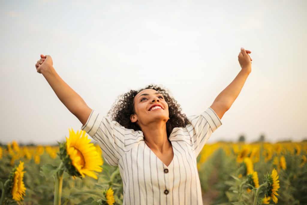 Mulher com os braços abertos, sorrindo e olhando para cima, em um campo de girassóis.