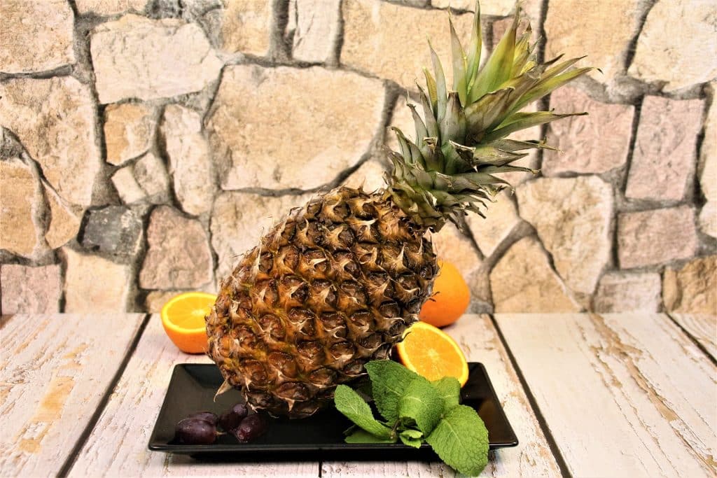 Imagem de um abacaxi. Ele está inteiro e sobre uma travessa de porcelana na cor preta, decorada com laranjas cortadas ao meio de folhas de hortelã.