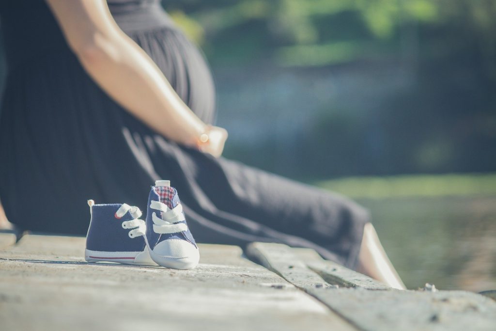 Imagem de uma mulher grávida. Ela veste um vestido preto e segura com suas mãos a barriga. Ela está sentada em um banco de concreto. Ao lado, um tênis azul de récem nascido.
