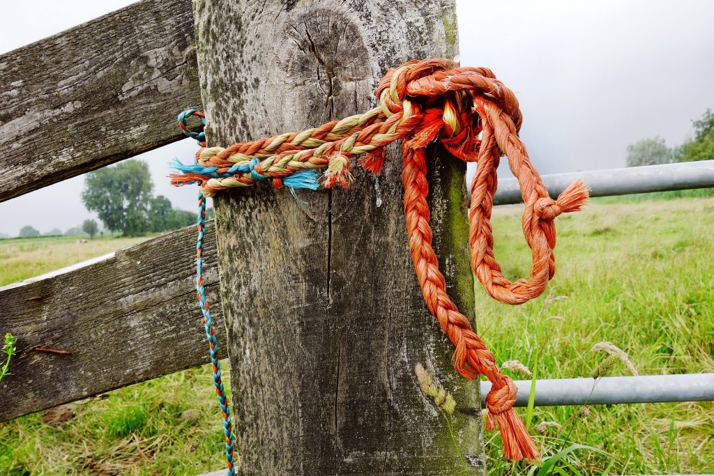 Imagem de uma corda alaranjada com um nó segurando a porteira de uma cerca de madeira.
