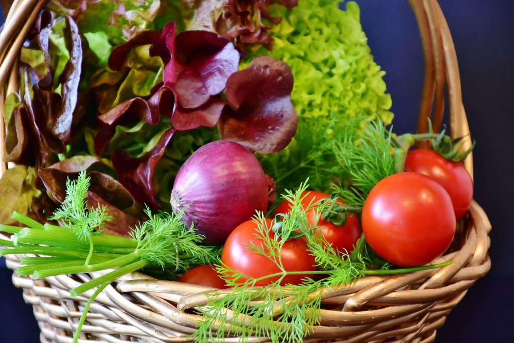 Imagem de um cesta de vime grande repleta de alimentos naturais e fotoprotetores como: verguras, legumes e ervas diversas.
