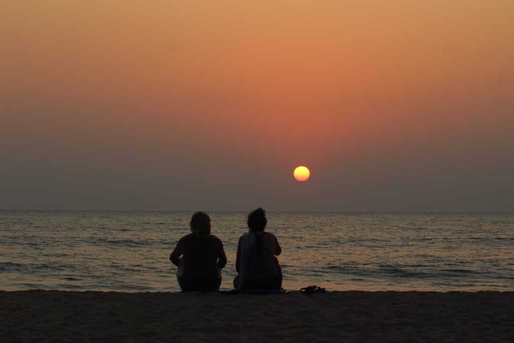 Imagem da silhueta de um casal homem e mulher sentandos na areia de uma praia assintindo o por do sol.
