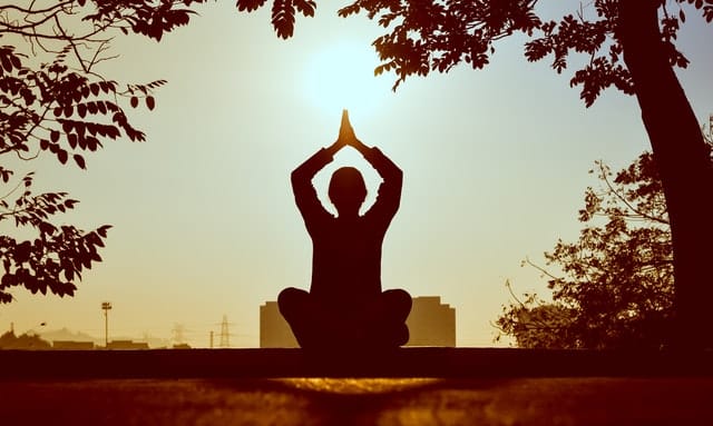 Silhueta de pessoa meditando, sentada e com os braços para cima, unindo as palmas das mãos.