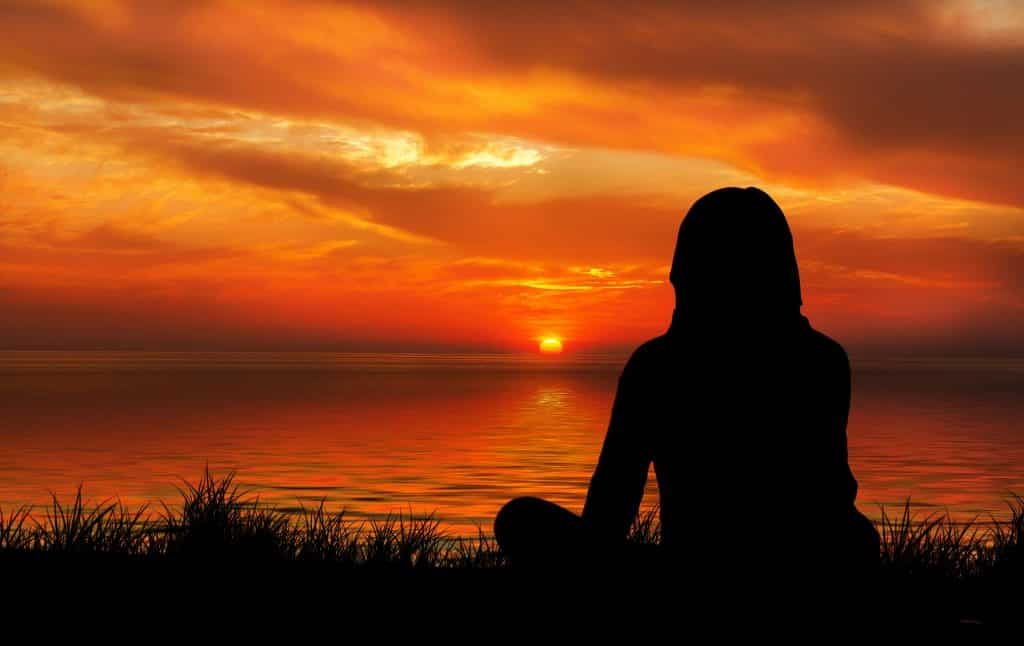 Imagem de uma mulher meditando em frente ao mar e apreciando um lindo por do sol.