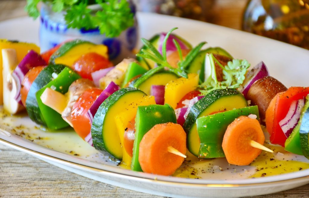 Imagem de um prato branco de porcelana repleto de lindos e saborosos espetos veganos feitos com cenoura, abobrinha, pimetão, tomate, cebola roxa e alecrim.
