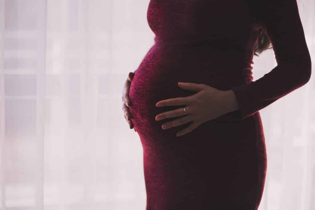 Imagem de uma mulher grávida de cabelos longos usando um vestido vermelho de frio. Ela está com as mãos sobre a barriga.