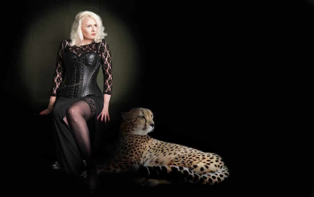 Mulher de  meia idade loura. Ela veste um lindo vestido preto com uma fenda lateral. Ela está sentada sobre um banquinho. Ao lado dela um lindo leopardo.
