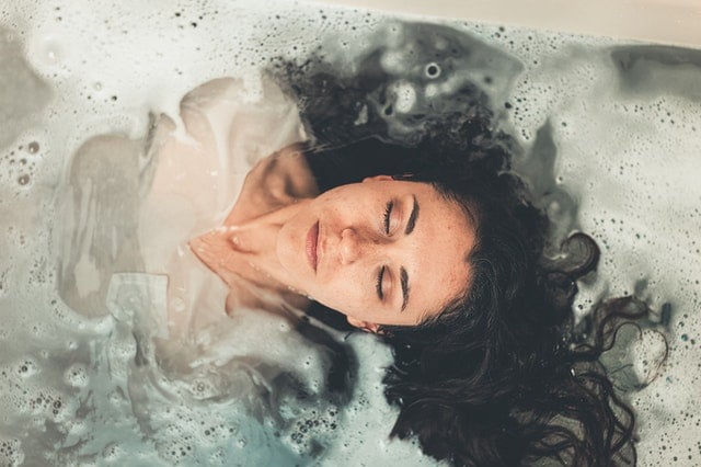 Mulher deitada em banheira com espumas em volta da cabeça vista do alto
