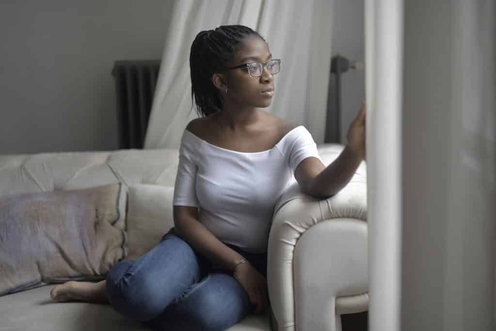 Mulher sentada no sofá de casa, olhando pela janela com expressão pensativa.
