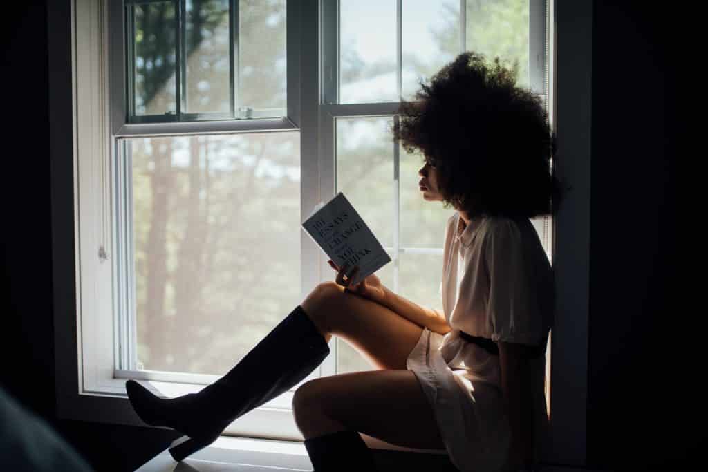 Mulher de botas e vestido sentada na janela, lendo um livro.