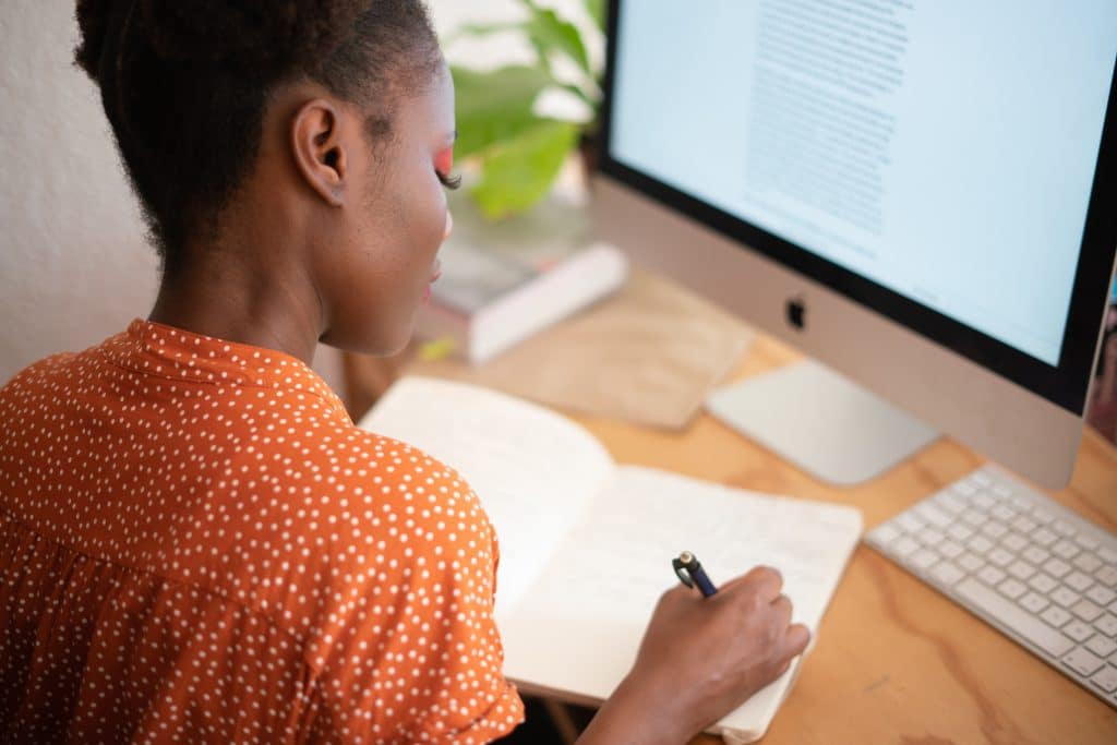 Mulher escrevendo em um caderno sobre uma mesa com um computador.