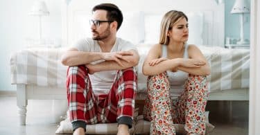 Homem e mulher usando pijamas, sentados lado a lado no chão, em frente a uma cama. Ambos olham para o lado oposto ao outro.