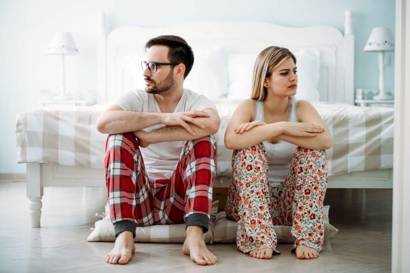 Homem e mulher usando pijamas, sentados lado a lado no chão, em frente a uma cama. Ambos olham para o lado oposto ao outro.