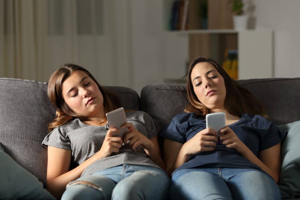 Duas mulheres sentadas em um sofá, relaxadas, e usando seus celulares.